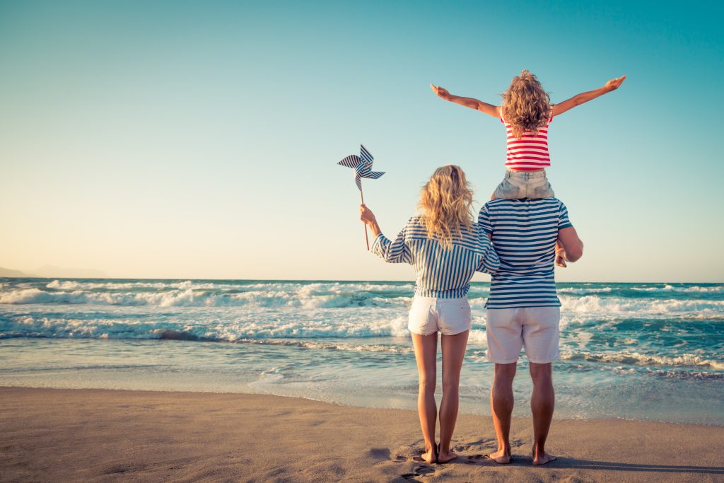 Das Foto zeigt eine Familie aus Vater, Mutter und Kind am Strand stehen und ins Meer gucken. Das Kind sitzt auf den Schultern des Vaters. Die Mutter hält ein Windrad in ihrer linken Hand. Sie haben rechtzeitig ihren Sommerurlaub gebucht.